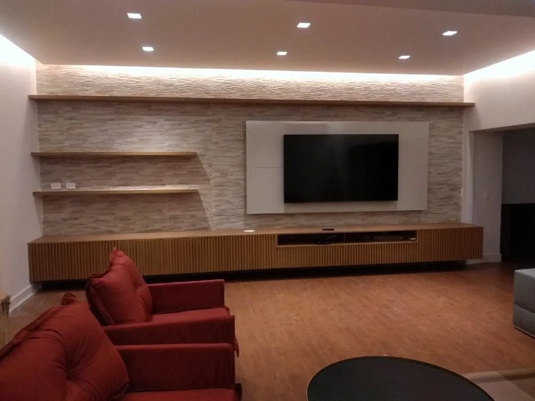 Sala com painel de TV em pedra, rack suspenso ripado em madeira e poltronas vermelhas