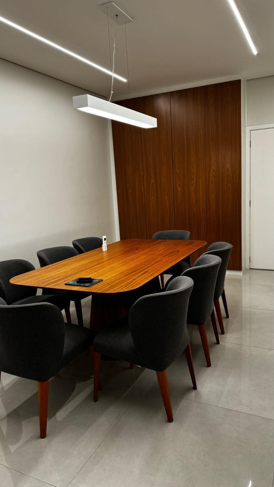 Sala de reunião com mesa de madeira, cadeiras cinza e painel em nogueira